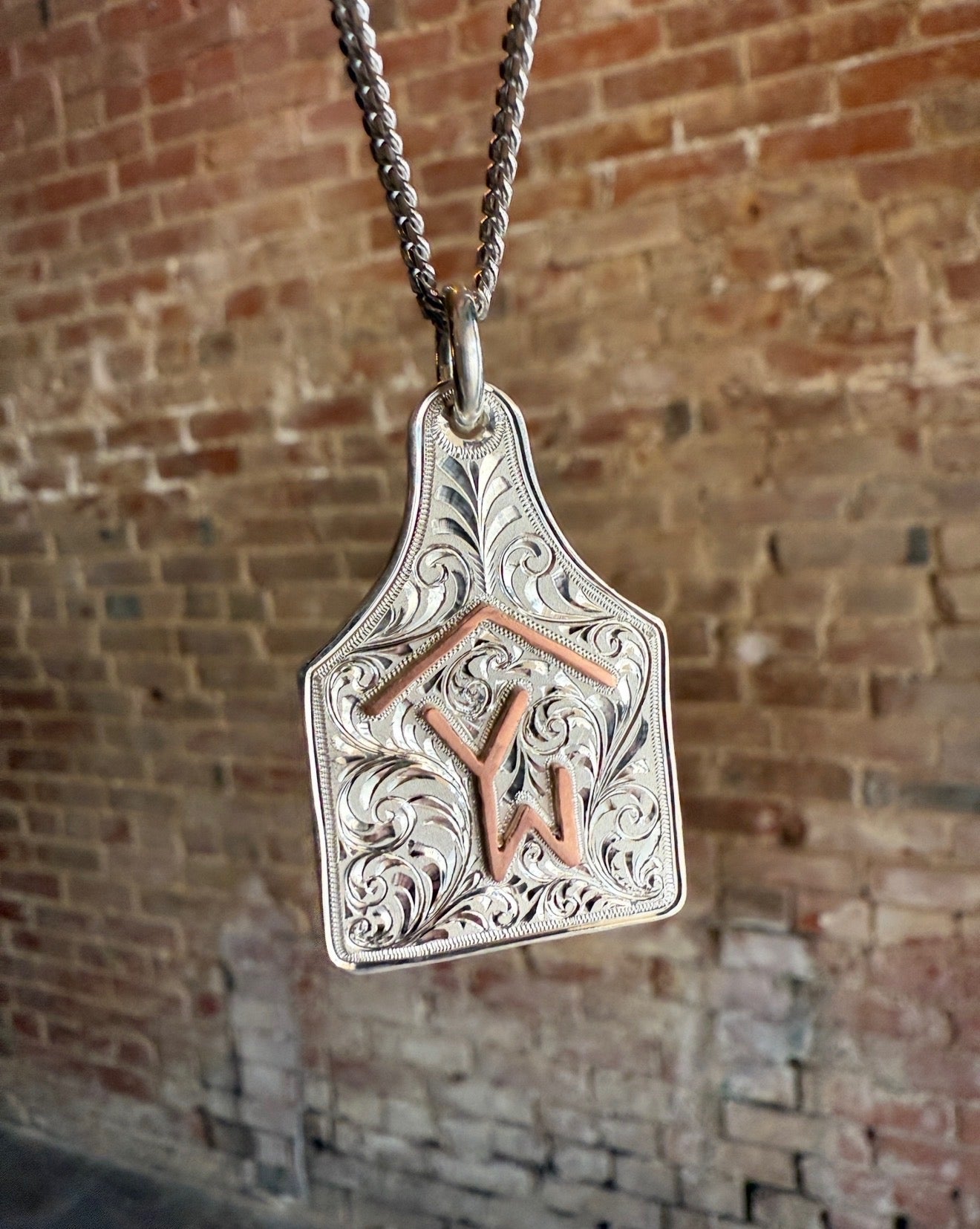 Silver pendant with engraved design and 'YW' initials against a brick wall background